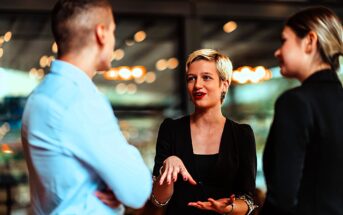 Three people are engaged in a conversation indoors. A woman with short blonde hair is speaking and gesturing with her hands while a man and woman listen attentively. The background features blurred lights and glass walls.