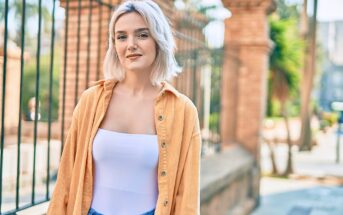 A woman with short blonde hair smiles slightly while standing outdoors. She wears an orange shirt over a white top and blue jeans. There are wrought iron fences and stone columns in the background. The setting appears to be sunny.