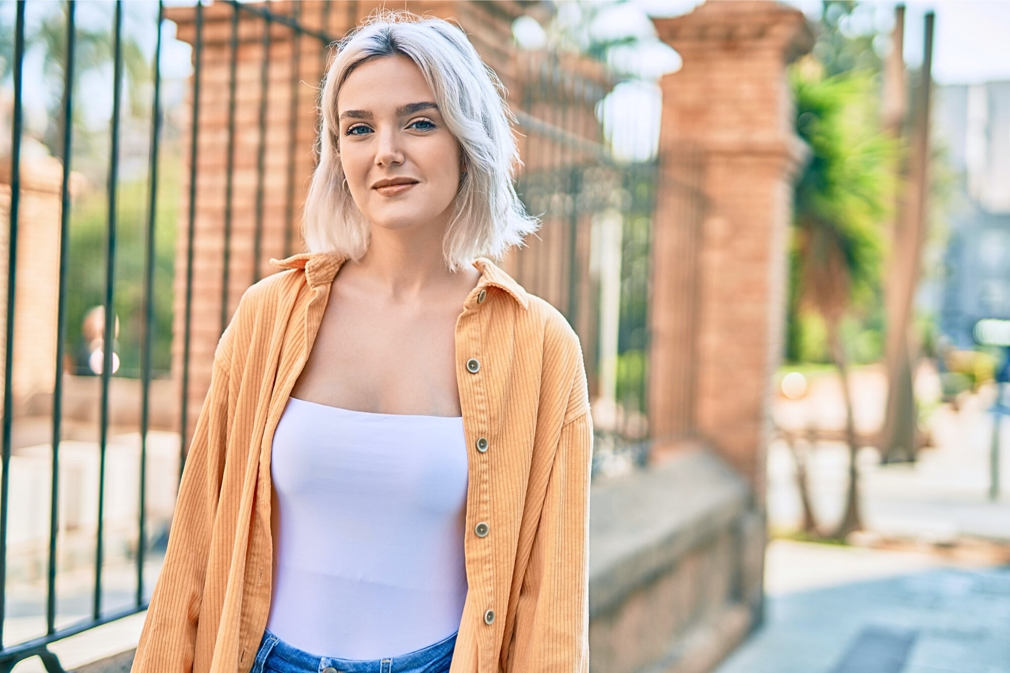 A woman with short blonde hair smiles slightly while standing outdoors. She wears an orange shirt over a white top and blue jeans. There are wrought iron fences and stone columns in the background. The setting appears to be sunny.