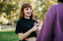 A woman with red hair and a black shirt gestures with her hands while talking to someone in a purple shirt, outdoors in a park setting. The background is sunny with green trees and grass.