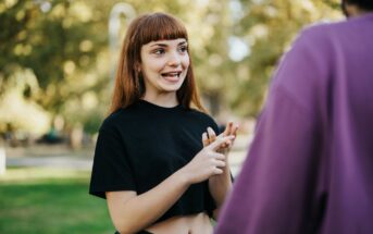 A woman with red hair and a black shirt gestures with her hands while talking to someone in a purple shirt, outdoors in a park setting. The background is sunny with green trees and grass.