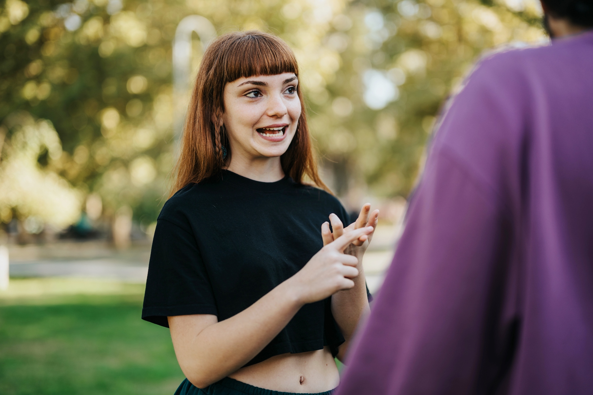 A woman with red hair and a black shirt gestures with her hands while talking to someone in a purple shirt, outdoors in a park setting. The background is sunny with green trees and grass.