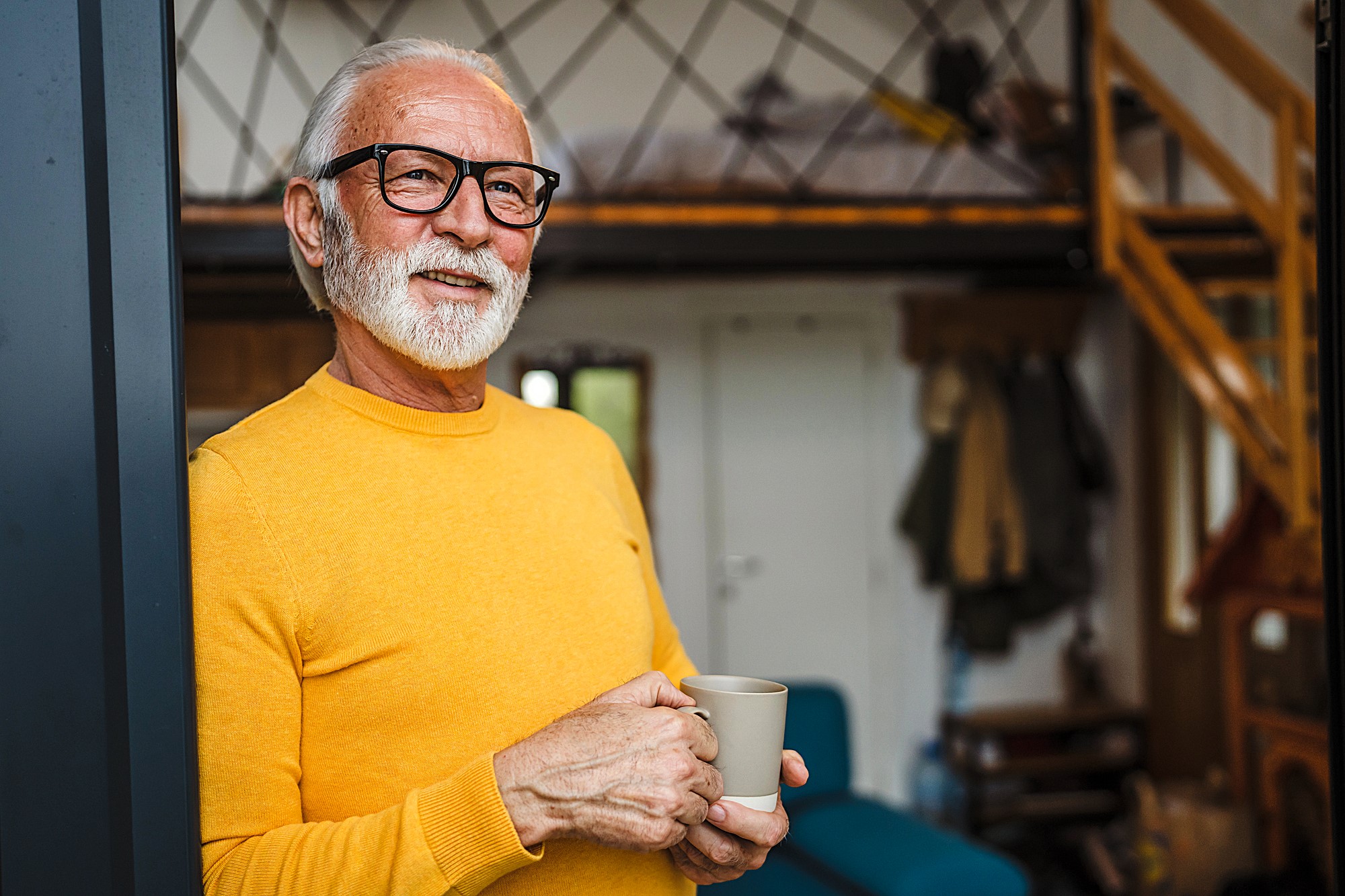 An older man with white hair and a beard is smiling while holding a mug. He is wearing a bright yellow sweater and glasses. The background shows a cozy, loft-style interior with a staircase and a railing.