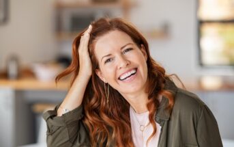 Smiling woman with long red hair, wearing a green shirt, gently touching her head. She's sitting in a bright kitchen with blurred background elements, including wooden shelves and countertops.