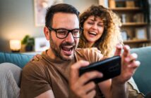 A man in glasses excitedly looks at a smartphone while a smiling woman with curly hair leans on his shoulder. They both appear to be enjoying something on the screen. The setting is a cozy living room with warm lighting.