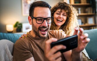 A man in glasses excitedly looks at a smartphone while a smiling woman with curly hair leans on his shoulder. They both appear to be enjoying something on the screen. The setting is a cozy living room with warm lighting.