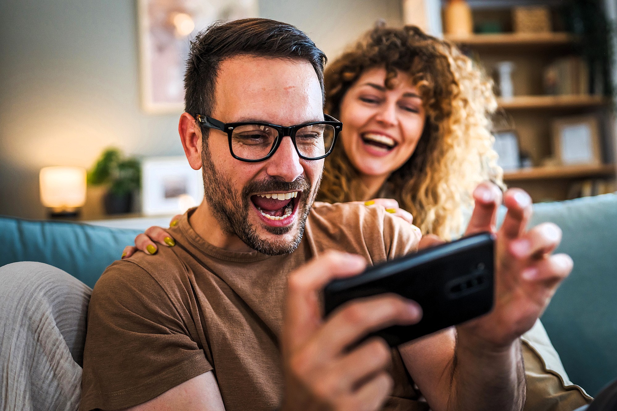 A man in glasses excitedly looks at a smartphone while a smiling woman with curly hair leans on his shoulder. They both appear to be enjoying something on the screen. The setting is a cozy living room with warm lighting.