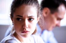 A woman with brown hair looks pensively off to the side, her expression serious. A man in the background appears blurred and focused on something else. The scene suggests a moment of contemplation or tension.