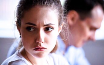 A woman with brown hair looks pensively off to the side, her expression serious. A man in the background appears blurred and focused on something else. The scene suggests a moment of contemplation or tension.