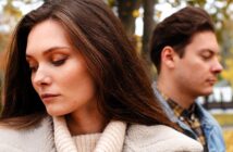 A woman and man stand back-to-back outdoors. The woman has long brown hair and is wearing a beige coat, looking down thoughtfully. The man, also with brown hair, wears a denim jacket and looks down. Trees with autumn leaves are in the background.