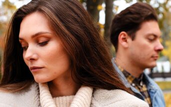 A woman and man stand back-to-back outdoors. The woman has long brown hair and is wearing a beige coat, looking down thoughtfully. The man, also with brown hair, wears a denim jacket and looks down. Trees with autumn leaves are in the background.