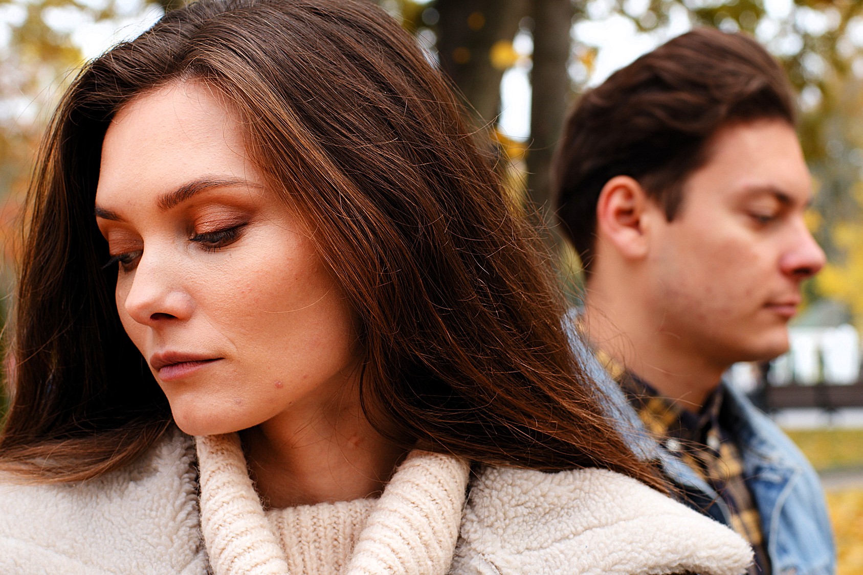 A woman and man stand back-to-back outdoors. The woman has long brown hair and is wearing a beige coat, looking down thoughtfully. The man, also with brown hair, wears a denim jacket and looks down. Trees with autumn leaves are in the background.