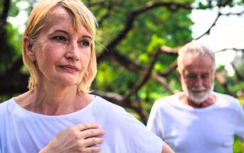 A middle-aged woman in a white shirt looks thoughtful while a man stands blurred in the background, also in a white shirt, in a lush green park setting.