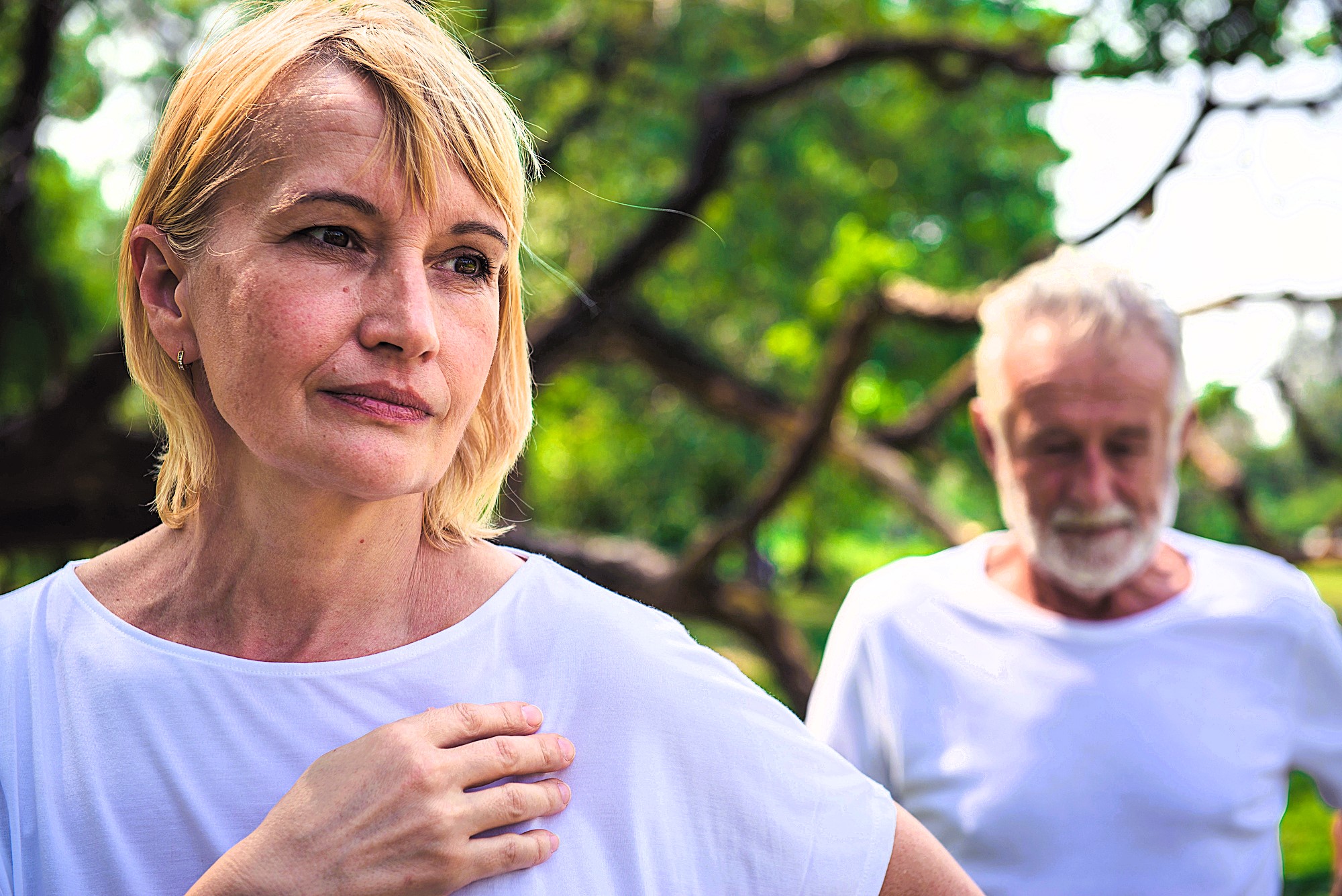 A middle-aged woman in a white shirt looks thoughtful while a man stands blurred in the background, also in a white shirt, in a lush green park setting.