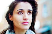 A woman with long dark hair looks upward, appearing thoughtful. She is outside, with a blurred background of buildings. She wears a light-colored scarf and has a gentle expression.