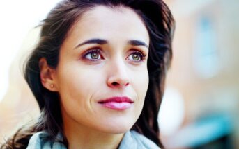 A woman with long dark hair looks upward, appearing thoughtful. She is outside, with a blurred background of buildings. She wears a light-colored scarf and has a gentle expression.