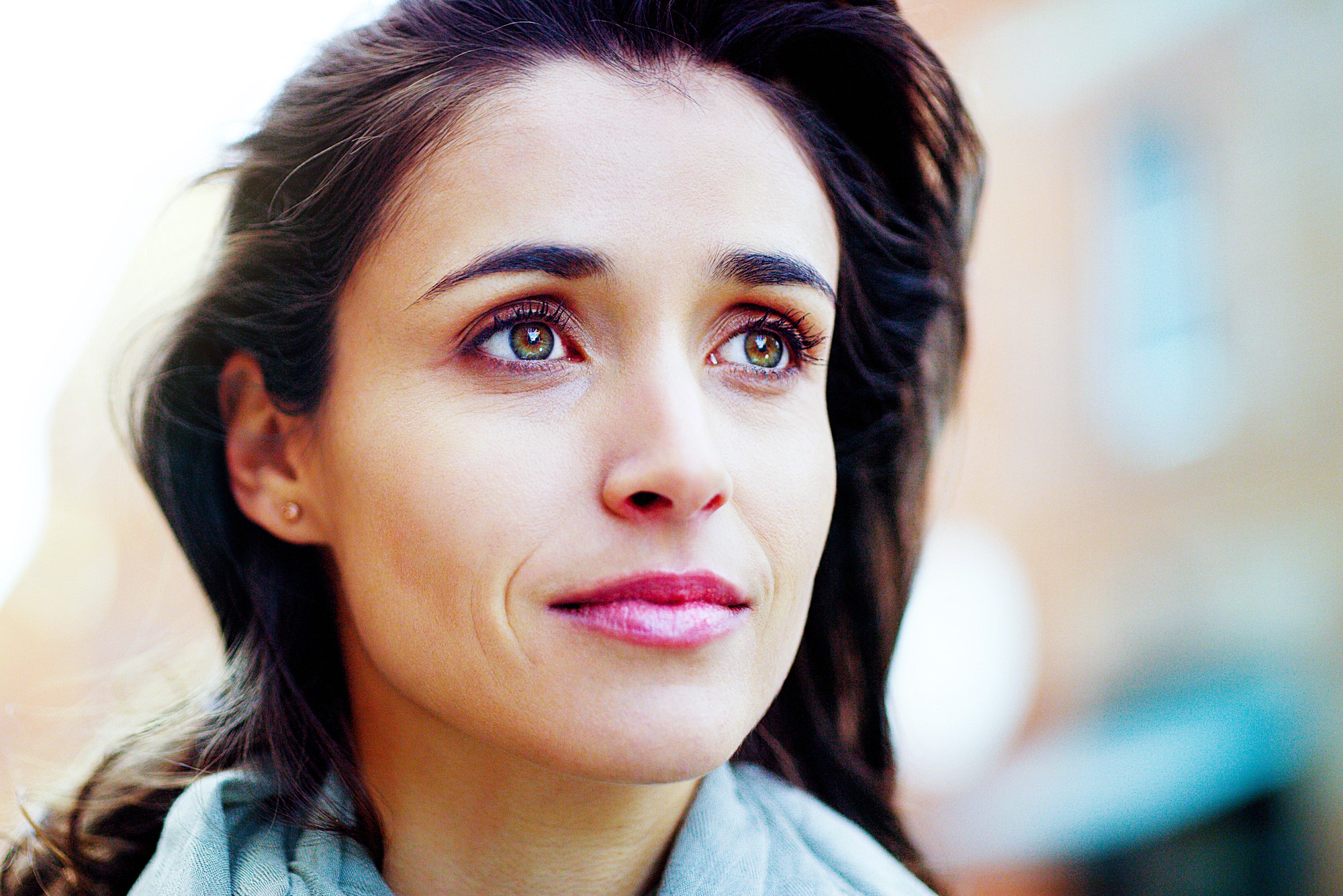 A woman with long dark hair looks upward, appearing thoughtful. She is outside, with a blurred background of buildings. She wears a light-colored scarf and has a gentle expression.