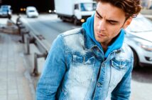 A man in a blue hoodie and denim jacket stands on a city street. He looks to the side with a pensive expression. Cars drive by on the road nearby.