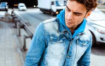 A man in a blue hoodie and denim jacket stands on a city street. He looks to the side with a pensive expression. Cars drive by on the road nearby.