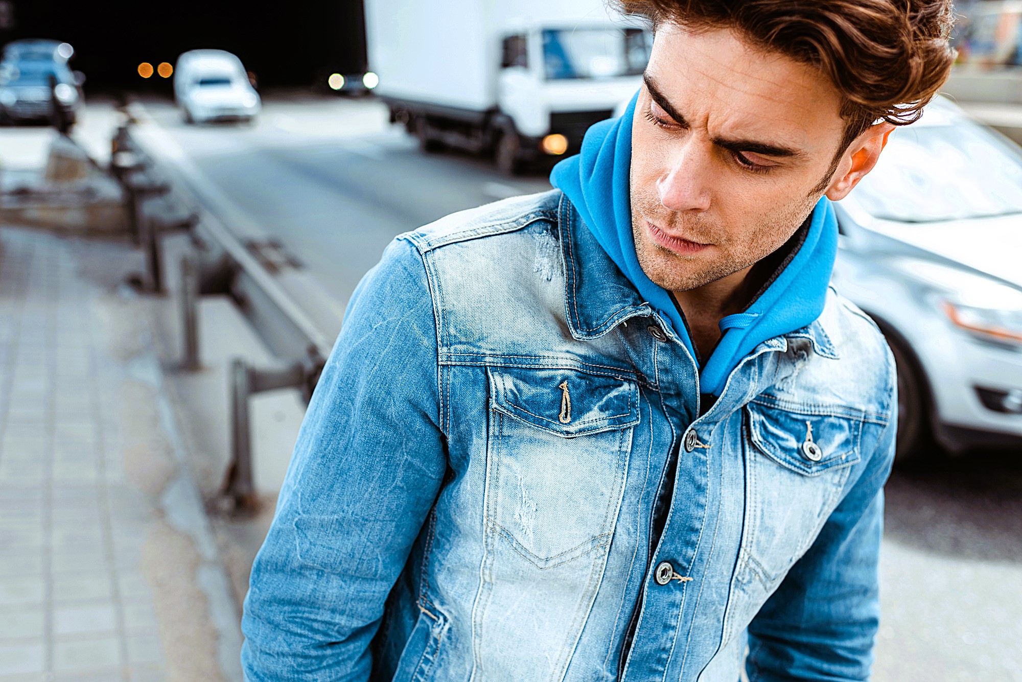 A man in a blue hoodie and denim jacket stands on a city street. He looks to the side with a pensive expression. Cars drive by on the road nearby.