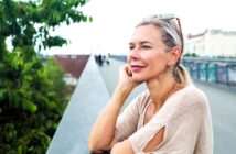 A woman with blonde hair and sunglasses on her head leans on a railing, looking thoughtfully into the distance. She is wearing a beige top with cutout sleeves. Green trees and blurred buildings are visible in the background.