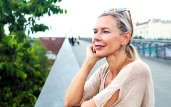 A woman with blonde hair and sunglasses on her head leans on a railing, looking thoughtfully into the distance. She is wearing a beige top with cutout sleeves. Green trees and blurred buildings are visible in the background.