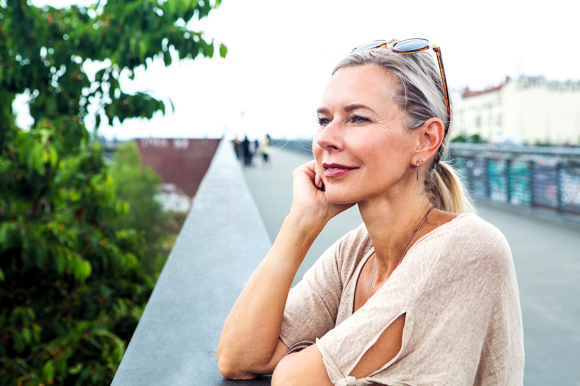 A woman with blonde hair and sunglasses on her head leans on a railing, looking thoughtfully into the distance. She is wearing a beige top with cutout sleeves. Green trees and blurred buildings are visible in the background.