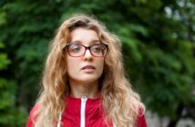 A woman with long, wavy hair and red-framed glasses is wearing a red jacket. She stands in front of a blurred green background, suggesting an outdoor setting with trees or foliage.