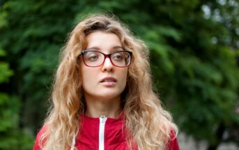 A woman with long, wavy hair and red-framed glasses is wearing a red jacket. She stands in front of a blurred green background, suggesting an outdoor setting with trees or foliage.