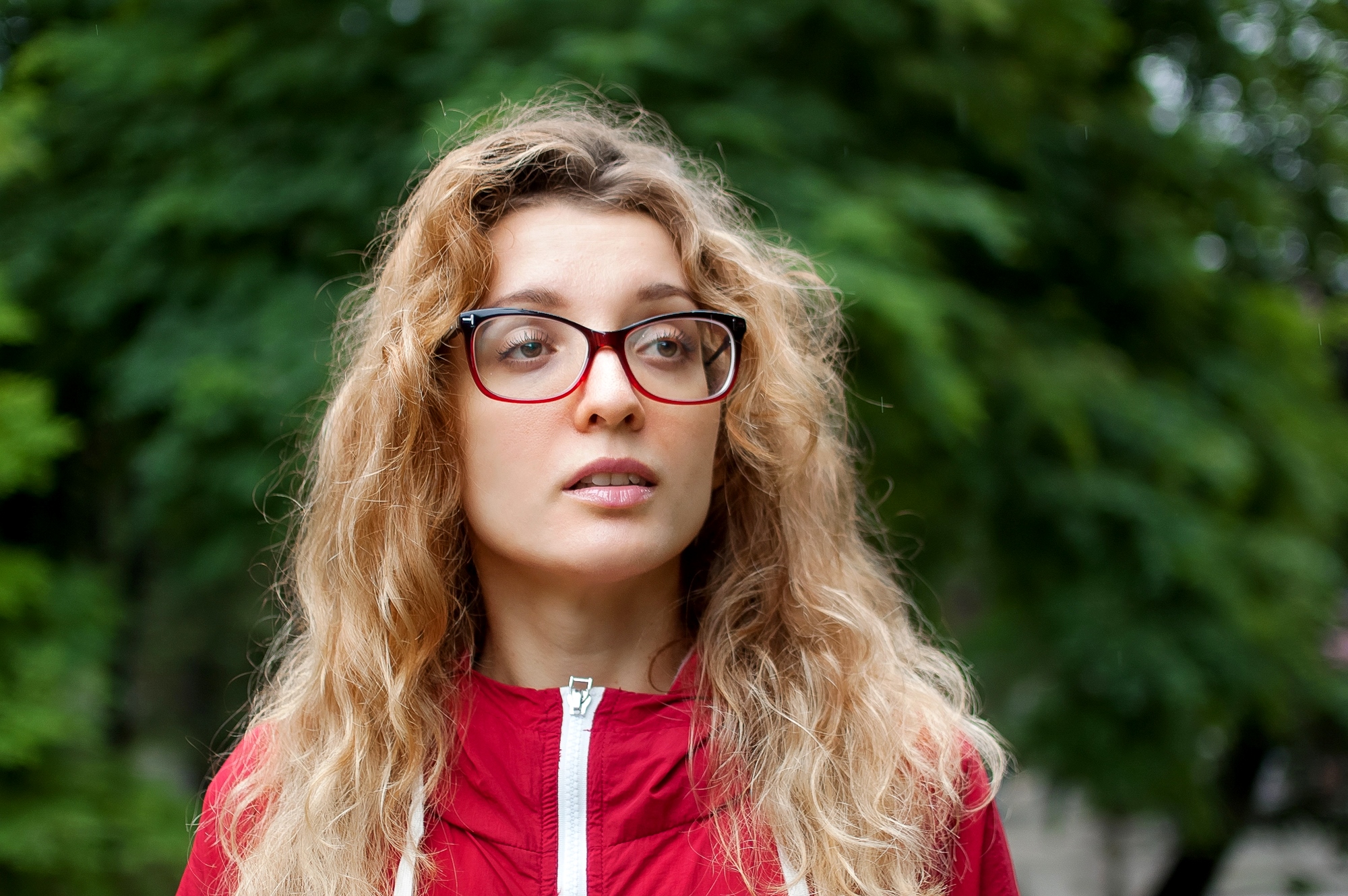 A woman with long, wavy hair and red-framed glasses is wearing a red jacket. She stands in front of a blurred green background, suggesting an outdoor setting with trees or foliage.