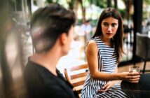 A woman in a striped dress sits at a table holding a drink, looking at a person with short hair. They appear to be engaged in conversation at an outdoor café with greenery in the background.