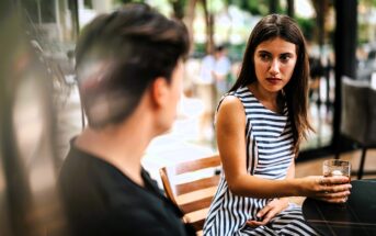 A woman in a striped dress sits at a table holding a drink, looking at a person with short hair. They appear to be engaged in conversation at an outdoor café with greenery in the background.