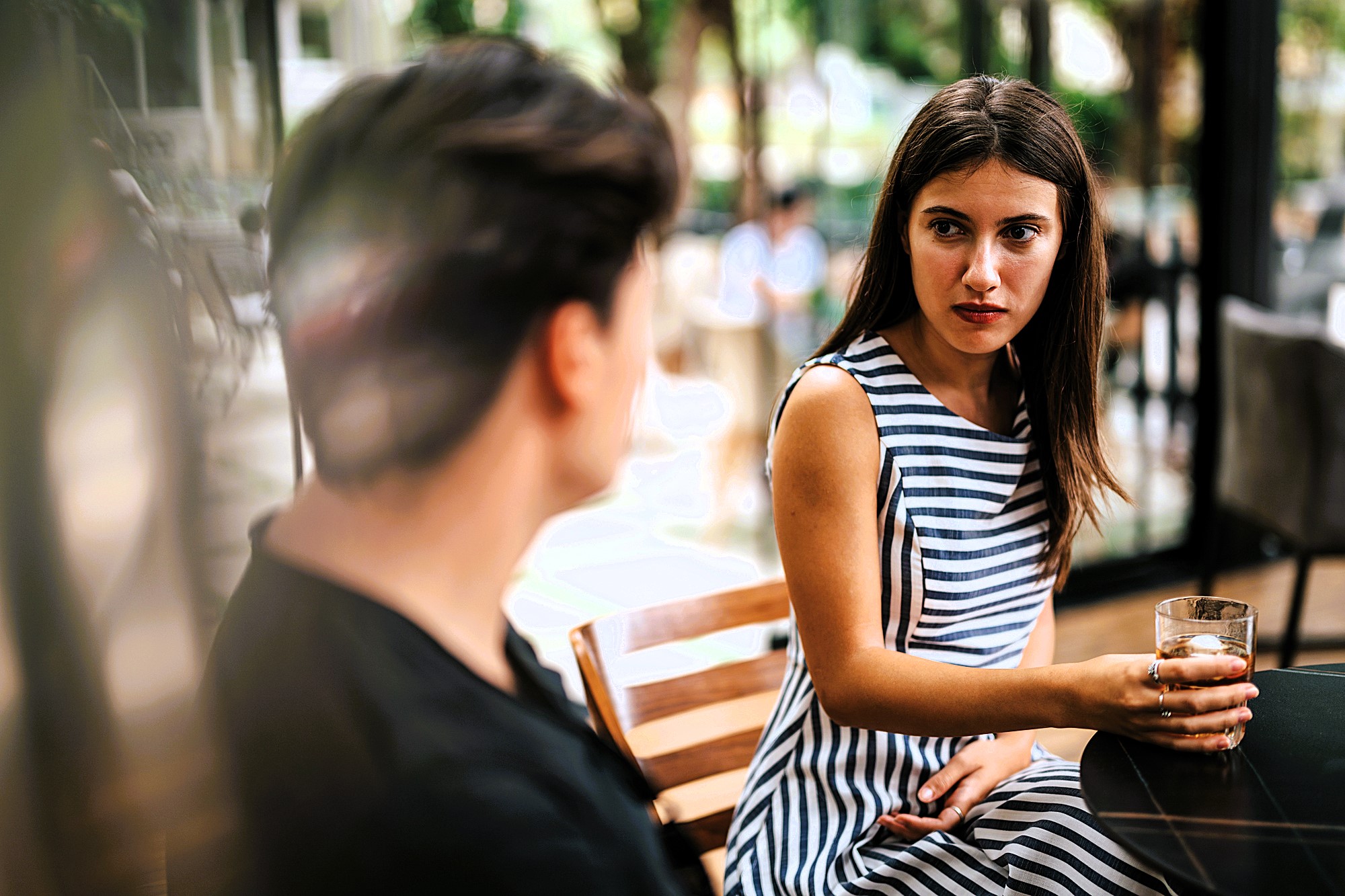 A woman in a striped dress sits at a table holding a drink, looking at a person with short hair. They appear to be engaged in conversation at an outdoor café with greenery in the background.