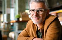 A man with gray hair and a beard, wearing glasses and a brown jacket, is smiling at the camera. The background features a cozy indoor setting with soft lighting and a plant on the table.