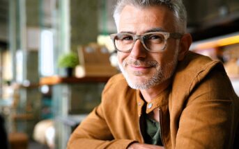 A man with gray hair and a beard, wearing glasses and a brown jacket, is smiling at the camera. The background features a cozy indoor setting with soft lighting and a plant on the table.