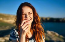 A woman with long red hair and a striped shirt is laughing joyfully outdoors. She stands near a body of water with a sunlit landscape in the background.