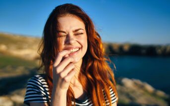 A woman with long red hair and a striped shirt is laughing joyfully outdoors. She stands near a body of water with a sunlit landscape in the background.