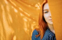 A person with long red hair stands partially obscured by a flowing orange fabric. They wear a blue polka-dotted shirt and are positioned against the bright backdrop, which casts soft shadows. The image captures a serene and contemplative mood.