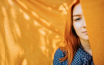 A person with long red hair stands partially obscured by a flowing orange fabric. They wear a blue polka-dotted shirt and are positioned against the bright backdrop, which casts soft shadows. The image captures a serene and contemplative mood.