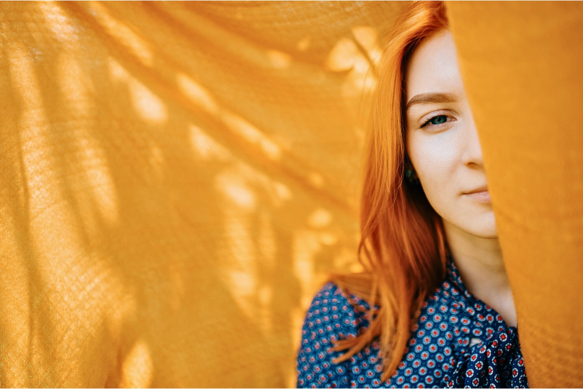 A person with long red hair stands partially obscured by a flowing orange fabric. They wear a blue polka-dotted shirt and are positioned against the bright backdrop, which casts soft shadows. The image captures a serene and contemplative mood.