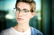 A person with short hair and glasses looks at the camera. They are wearing a white shirt and denim jacket. The background is blurry, with a mix of green and blue tones.