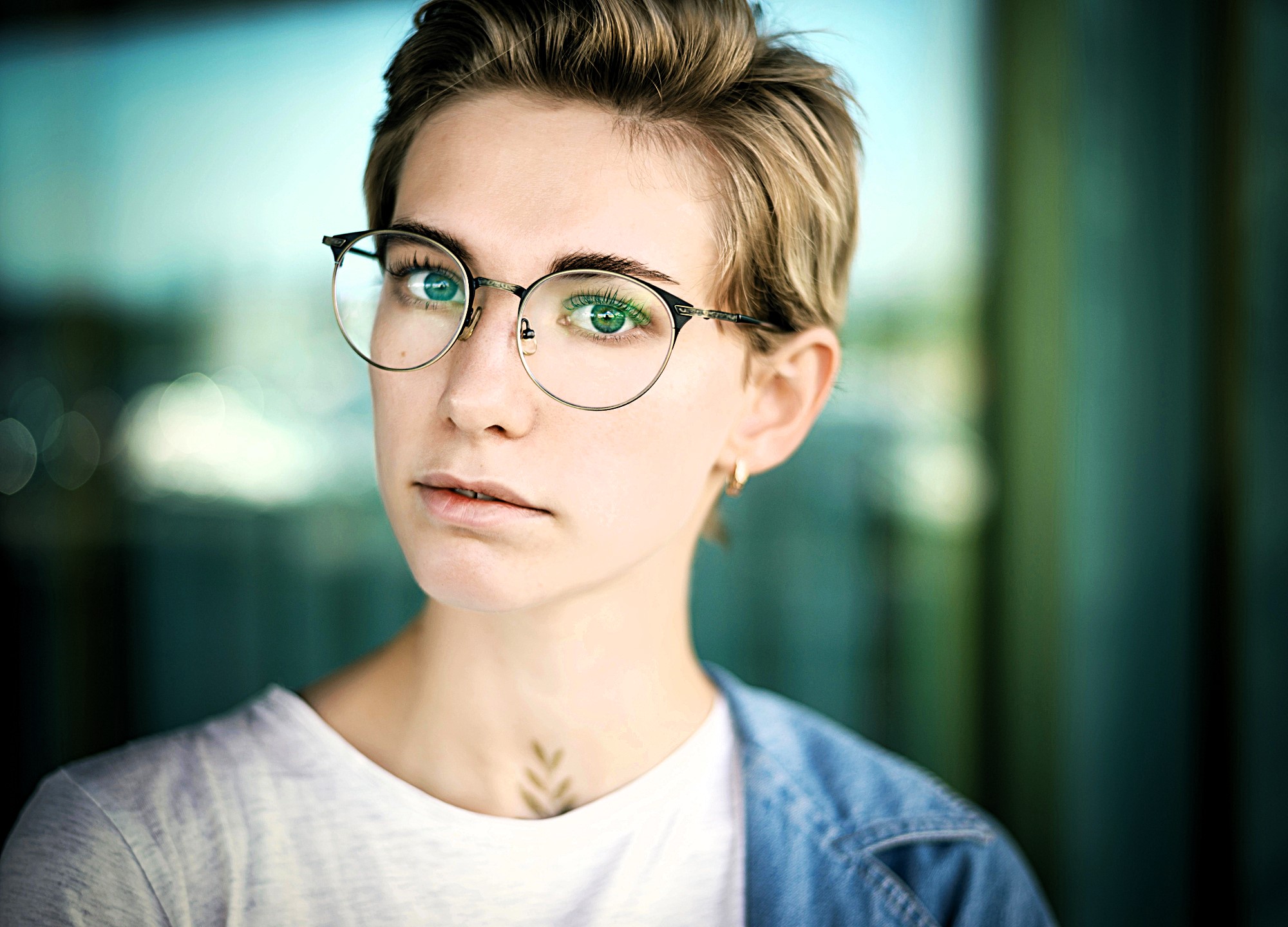 A person with short hair and glasses looks at the camera. They are wearing a white shirt and denim jacket. The background is blurry, with a mix of green and blue tones.