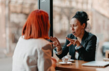 Two women sit across from each other at a wooden table in a café, engaging in conversation. One woman with red hair listens while the other gestures expressively. They each have a small cup of coffee in front of them.