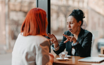 Two women sit across from each other at a wooden table in a café, engaging in conversation. One woman with red hair listens while the other gestures expressively. They each have a small cup of coffee in front of them.