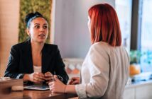 Two women engaged in conversation at a counter. One woman with red hair faces away; the other, with dark hair in a bun, listens attentively. The background includes a green wall and light wooden surfaces, creating a modern and inviting atmosphere.