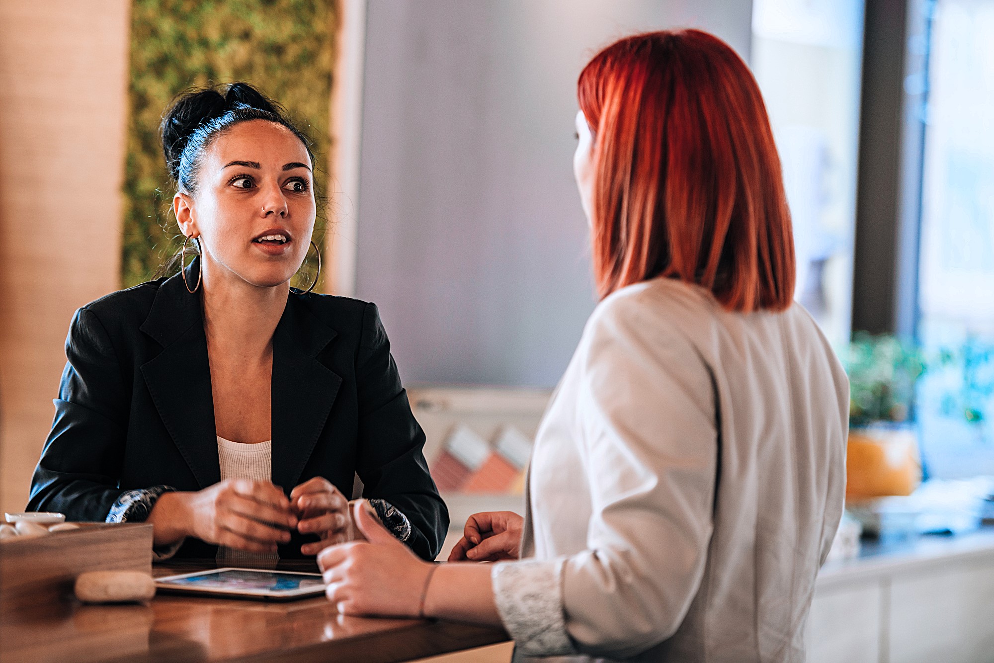 Two women engaged in conversation at a counter. One woman with red hair faces away; the other, with dark hair in a bun, listens attentively. The background includes a green wall and light wooden surfaces, creating a modern and inviting atmosphere.