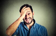 A man wearing a blue shirt with a distressed expression holds his forehead, appearing frustrated or in pain. The background is a textured gray wall.