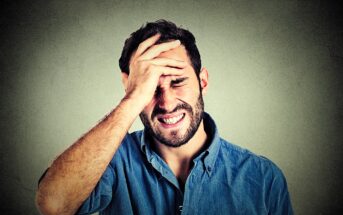 A man wearing a blue shirt with a distressed expression holds his forehead, appearing frustrated or in pain. The background is a textured gray wall.