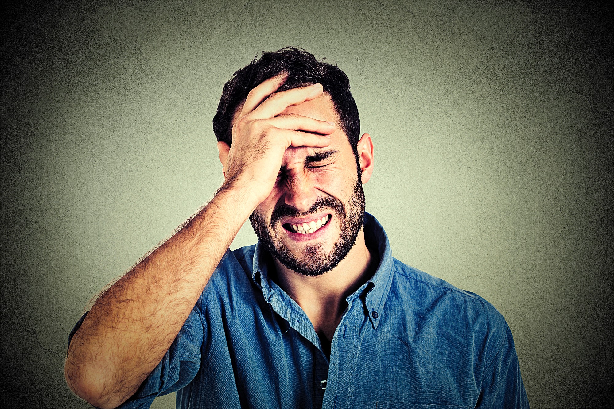 A man wearing a blue shirt with a distressed expression holds his forehead, appearing frustrated or in pain. The background is a textured gray wall.