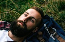 A bearded man lies on his back in the grass, resting his head on a blue backpack. He is wearing a plaid shirt over a white T-shirt and is looking directly at the camera.